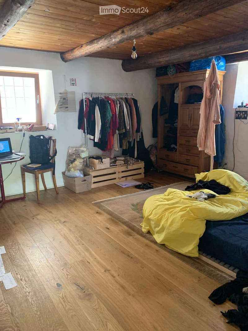 room with wooden floor, wooden beams, white walls, bed, chair, desk, wardrobe, clothes rack, storage boxes, laptop, bag