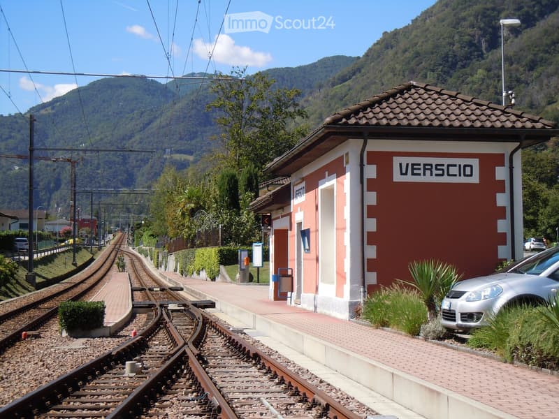 Railway station with a small building, platform, and tracks, surrounded by greenery and mountains