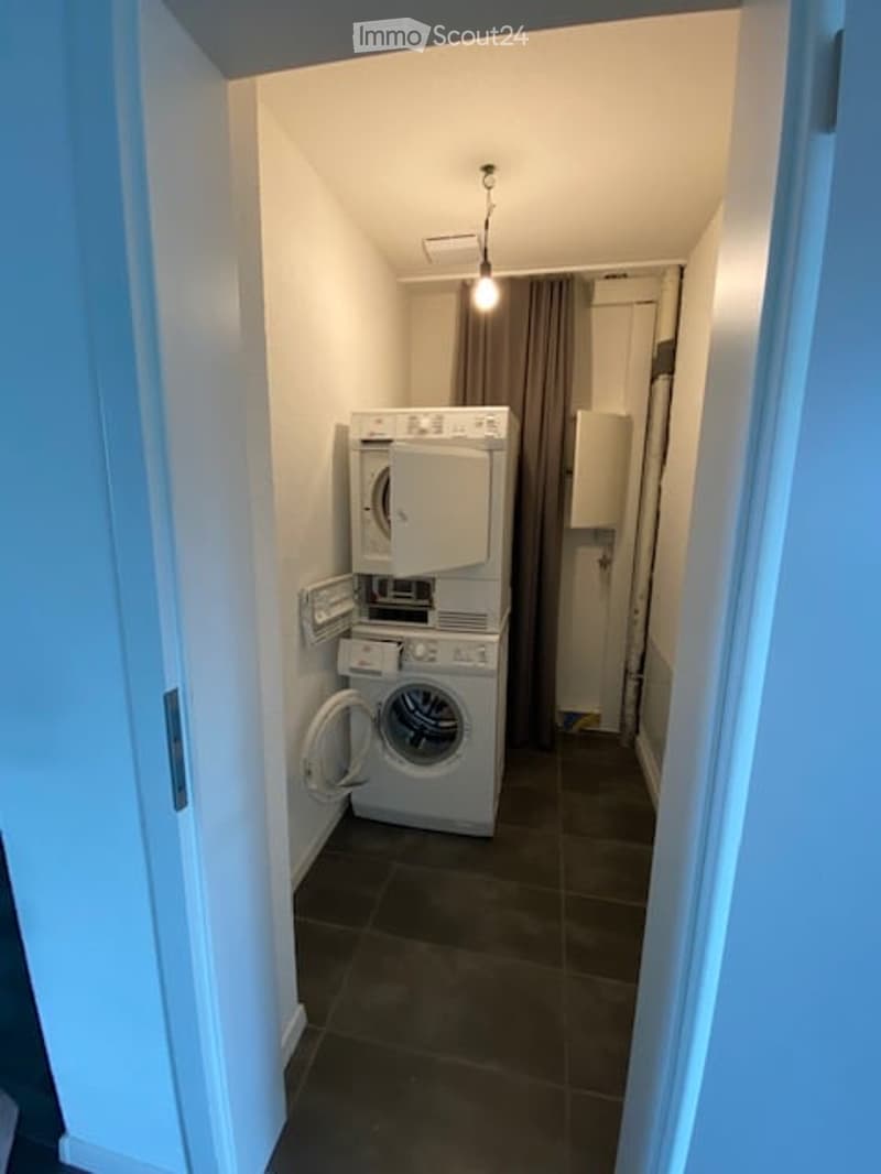 Furnished Room. Compact laundry room with stacked washing machine and dryer, dark tiled floor, white walls, and a hanging light fixture.