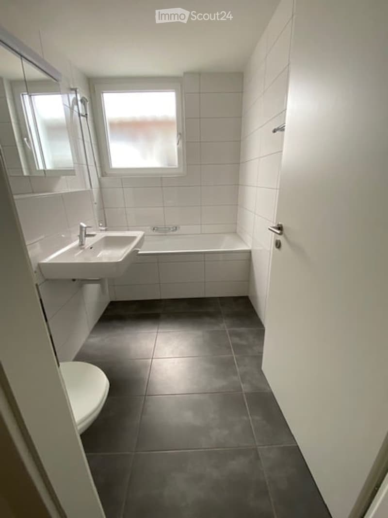 Bathroom. Modern bathroom with white tiled walls, dark grey tiled floor, a white sink, a bathtub, and a toilet. A window provides natural light, and a mirror is mounted on the wall above the sink.