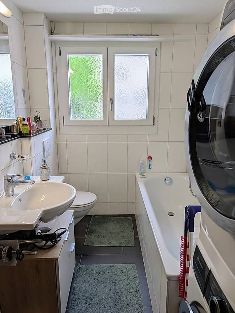 Bathroom. Modern bathroom with white tiles, a sink, toilet, bathtub, and a washing machine. The room features a window with frosted glass, a mirror above the sink, and a small shelf with toiletries.