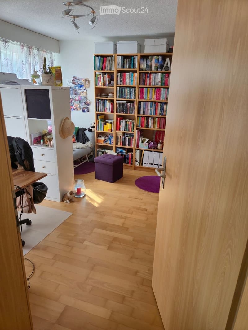Furnished Room. A cozy, well-lit room with wooden flooring, a large bookshelf filled with books, a small table with a chair, a purple ottoman, and a small dog near a lit candle. The room features a curtained window and a partially visible bed area with a