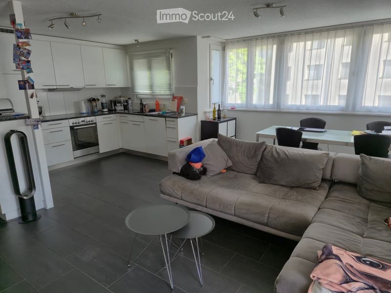Furnished Room. Modern living room with a grey sectional sofa, two round coffee tables, a dining table with chairs, and an open kitchen area with white cabinets, an oven, and a refrigerator.