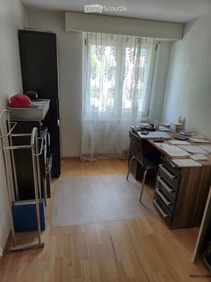 Furnished Room. A well-lit room with wooden flooring, a large window with sheer curtains, a desk with a laptop, chair, and various office supplies, a cabinet with hats and books, and a small rug in the center.
