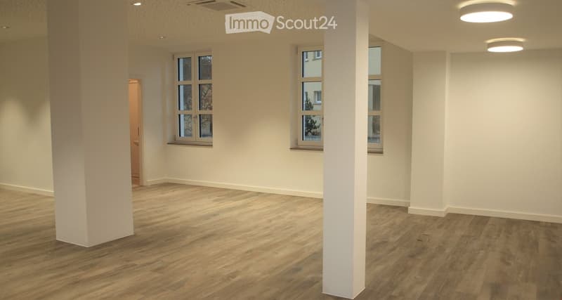 An empty room with wood flooring, three windows, two central columns, ceiling lights.