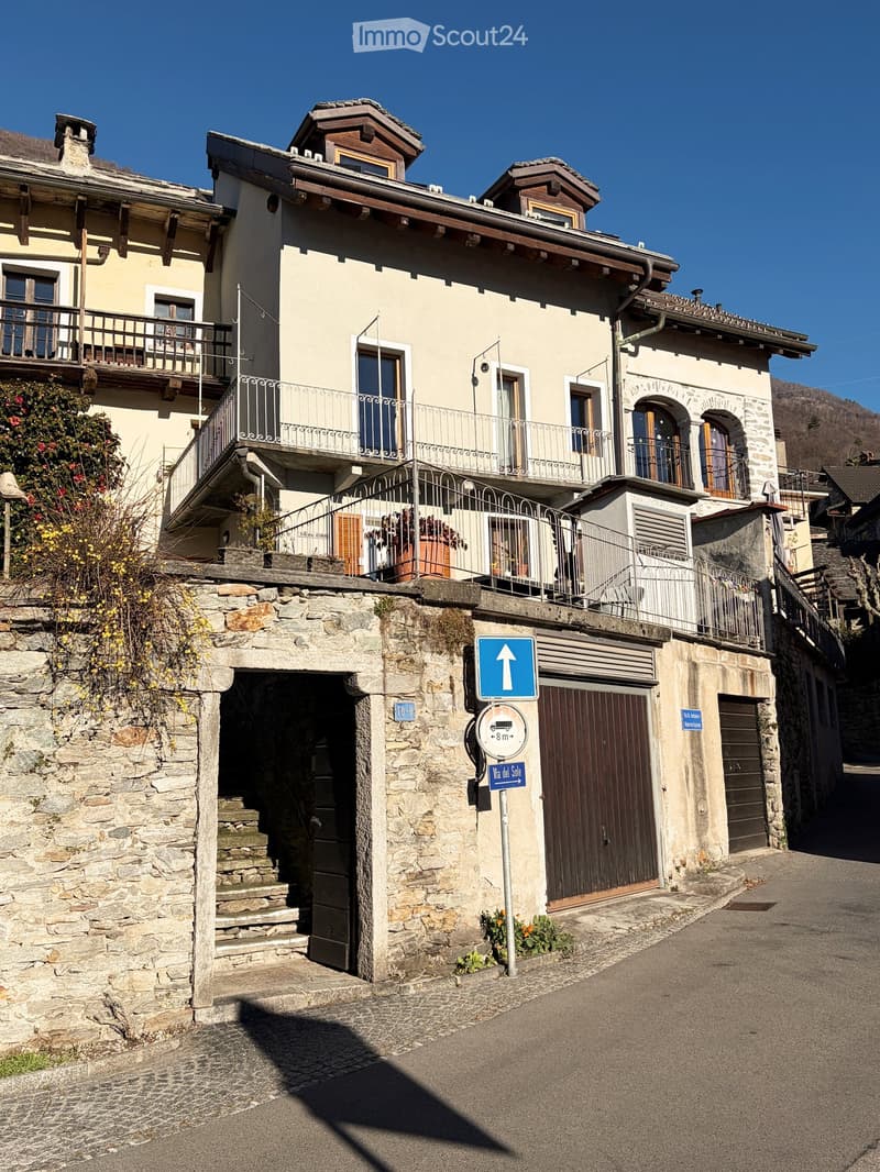 The building has multiple stories, exposed stonework on the lower portion, balconies with iron railings, stairs leading to a lower entrance, two garages with brown doors, and a blue sign indicating height restrictions.