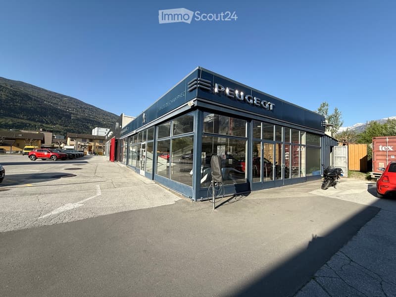 Building. A modern Peugeot car dealership with large glass windows and a flat roof, situated in a commercial area with visible cars and motorcycles.