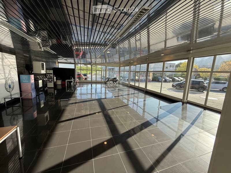 Corridor / Hallway. Modern, spacious hallway with large glass windows and a sleek, reflective tiled floor. The ceiling features a geometric, angular design with integrated lighting. Visible outside are parked cars and a green landscape.