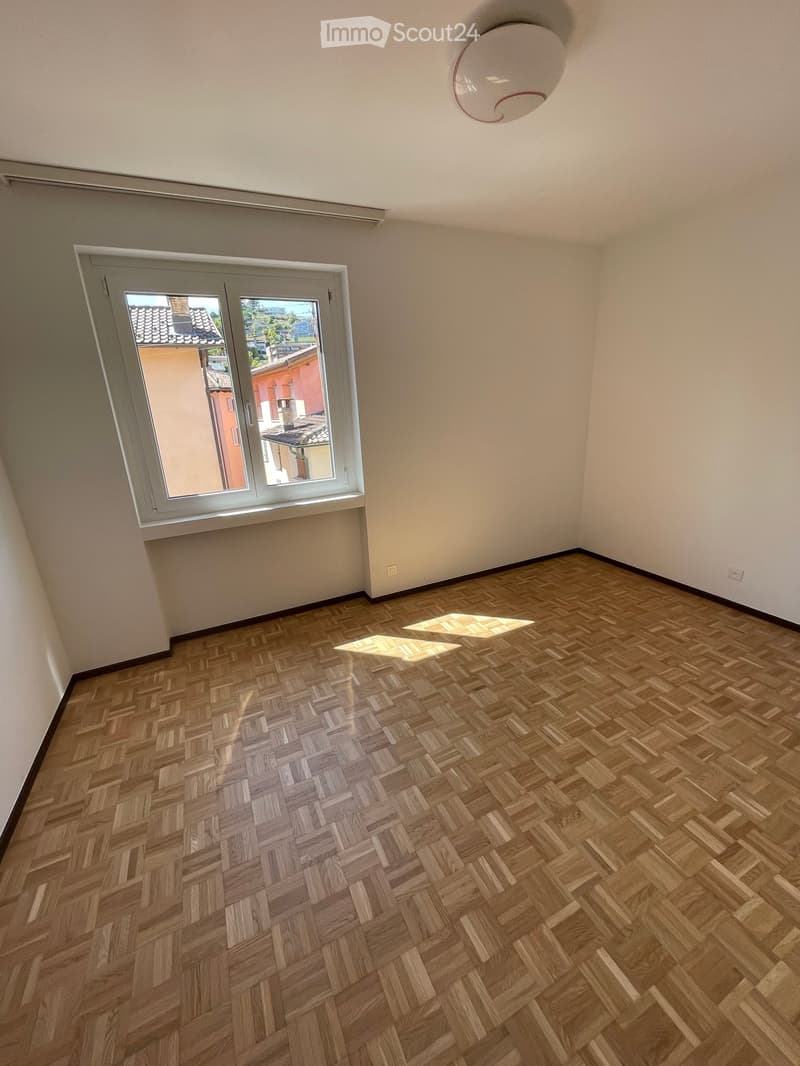 Unfurnished Room. Empty room with wooden herringbone flooring, white walls, and a large window with a view of the outside. The room has a modern ceiling light fixture.