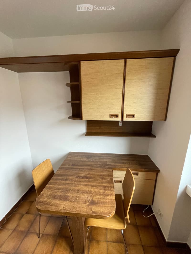 Furnished Room. A small, furnished room with a wooden table, two chairs, and built-in wooden cabinets with shelves above.