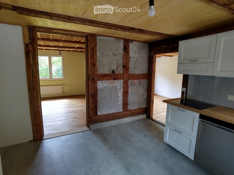 Unfurnished Room. An empty room with wooden beams on the ceiling, a stone wall, and a wooden floor. There are white cabinets and a countertop with a black stove top. A white radiator is mounted on the wall, and a window with a view of trees outside is on