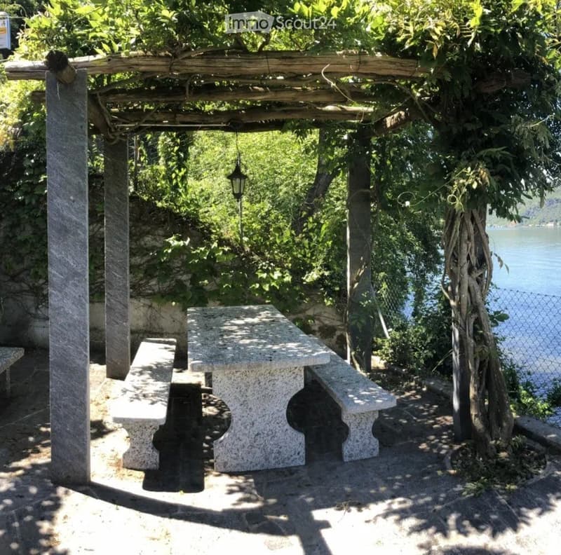 Garden / Outdoor Area. A shaded outdoor seating area with a stone table and benches under a wooden pergola, surrounded by greenery and overlooking a body of water.
