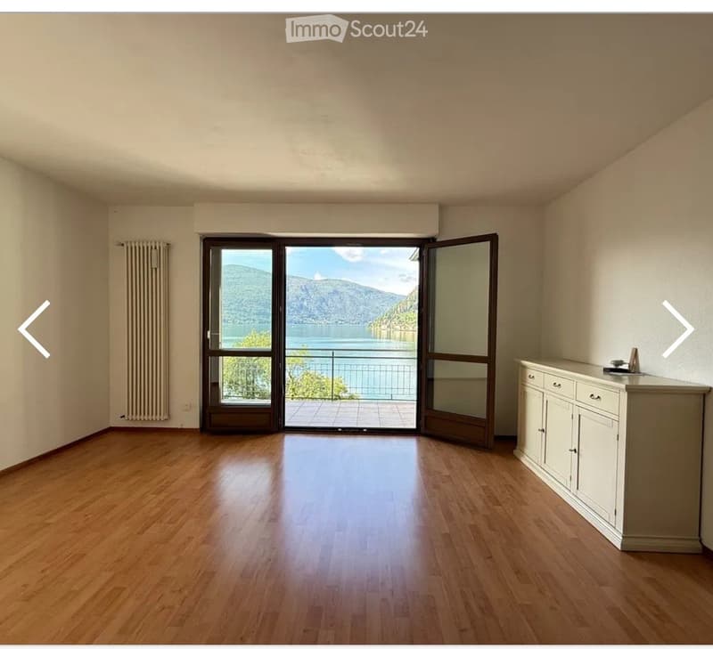 Unfurnished Room. Empty room with wooden flooring, large sliding glass doors leading to a balcony with a scenic lake and mountain view, and a small cabinet with drawers on the right side.