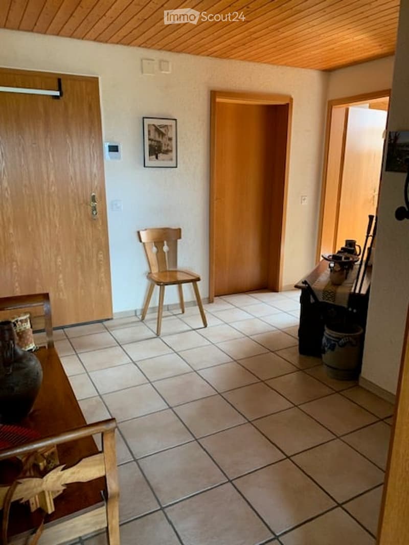 Furnished Room. A small, well-lit room with tiled flooring and wooden ceiling. Features include a wooden chair, a small table with a vase, and a picture frame on the wall.