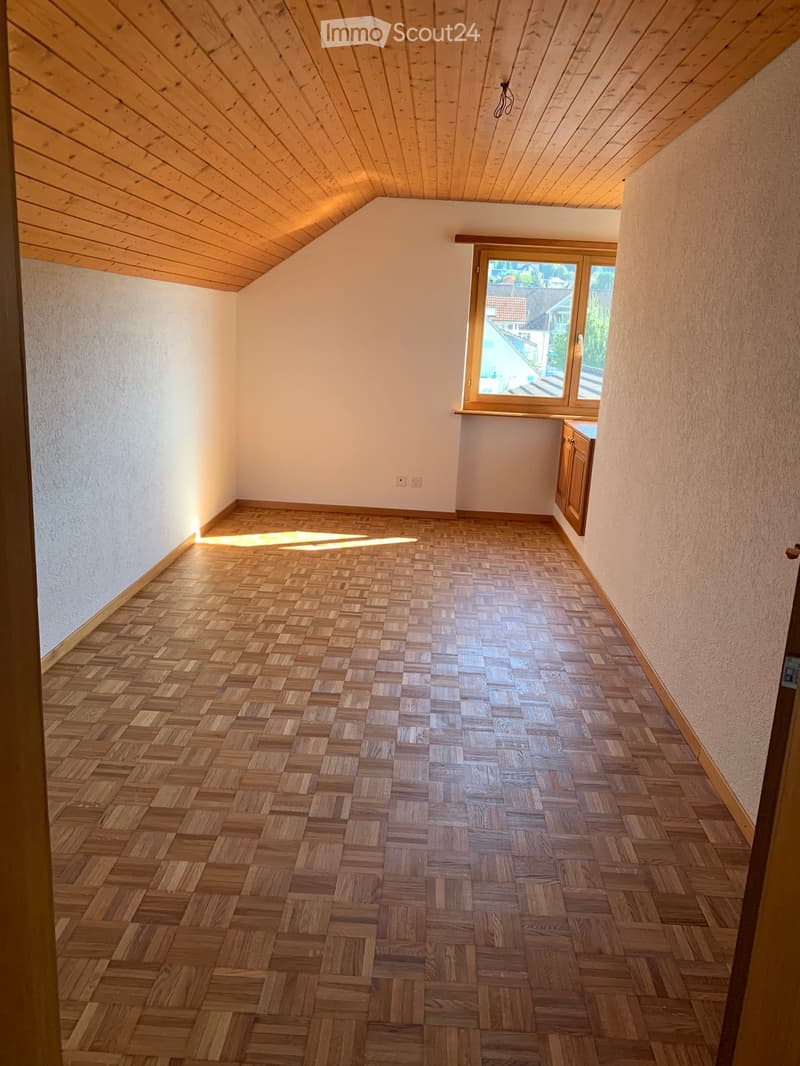 Unfurnished Room. Empty room with wooden herringbone flooring, white walls, and a slanted wooden ceiling. A window with wooden frame and a small cabinet are visible.
