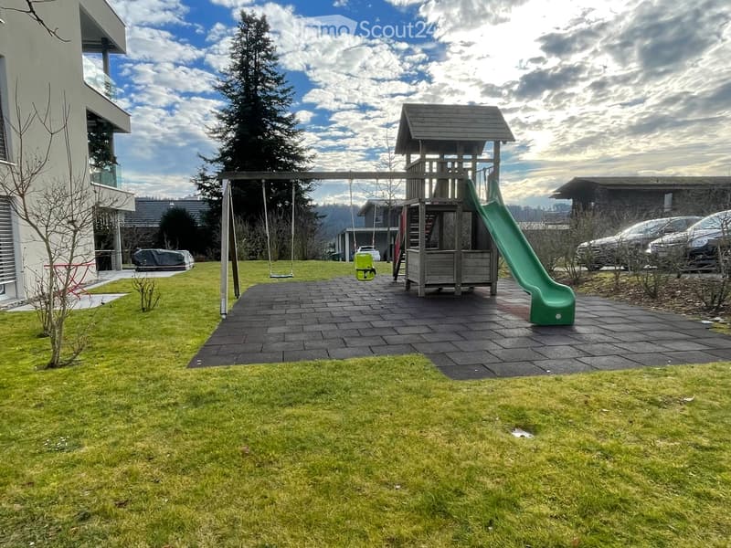 A playground featuring a green slide, two swings, a wooden playhouse, and a paved area with rubber tiles.