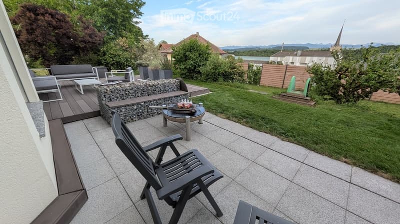 Outdoor patio with tiled flooring, lounge chairs, small table, and a view of a landscaped garden with a stone wall, barbecue, and surrounding greenery.
