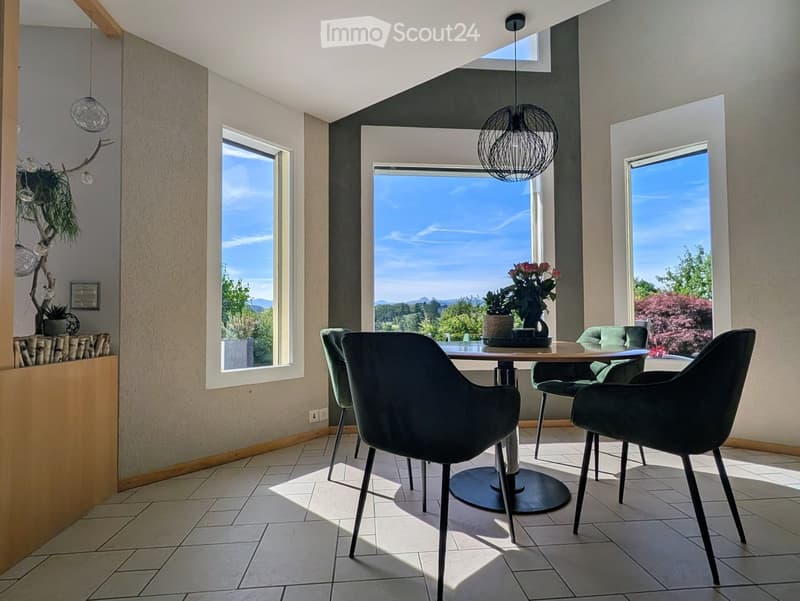 Modern dining room with large windows offering scenic views, a round dining table with four chairs, a flower vase on the table, and a stylish pendant light hanging from the ceiling.