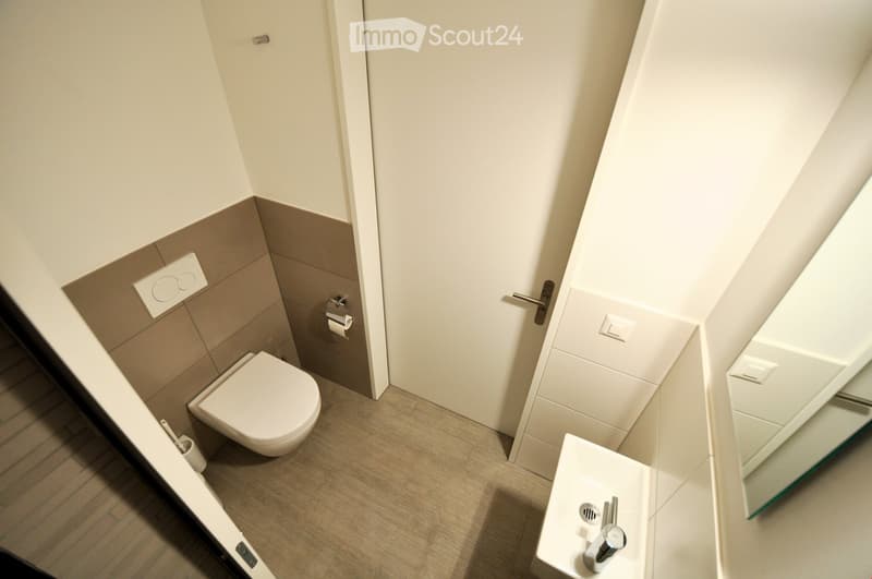 Bathroom. Modern bathroom with a toilet, sink, and mirror. The walls are painted in neutral tones with a grey accent wall. The floor is tiled in a light wood pattern.