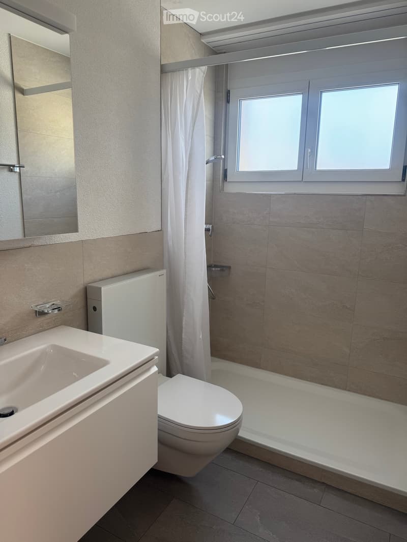 Bathroom. Modern bathroom with a white sink, toilet, mirror, and shower area with a curtain. Light-colored tiles and a window provide natural light.