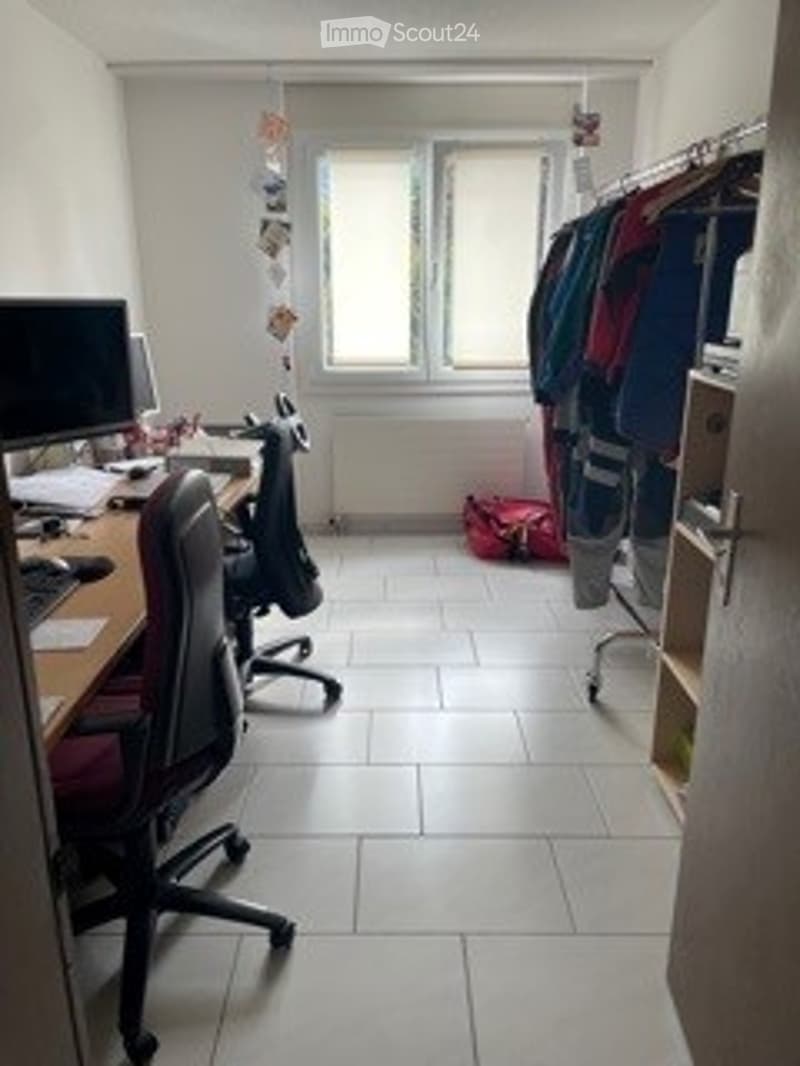 Furnished Room. A small, furnished room with a desk, computer setup, and a clothing rack. The room has white tiled flooring and large windows allowing natural light.