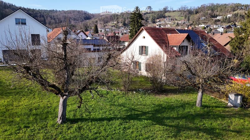House. Residential houses with red-tiled roofs and white walls, surrounded by green grass and trees in a hilly landscape.