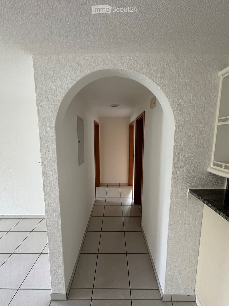 Corridor / Hallway. A narrow hallway with tiled flooring, white textured walls, and an arched entryway leading to another room. Wooden doors are visible on both sides.