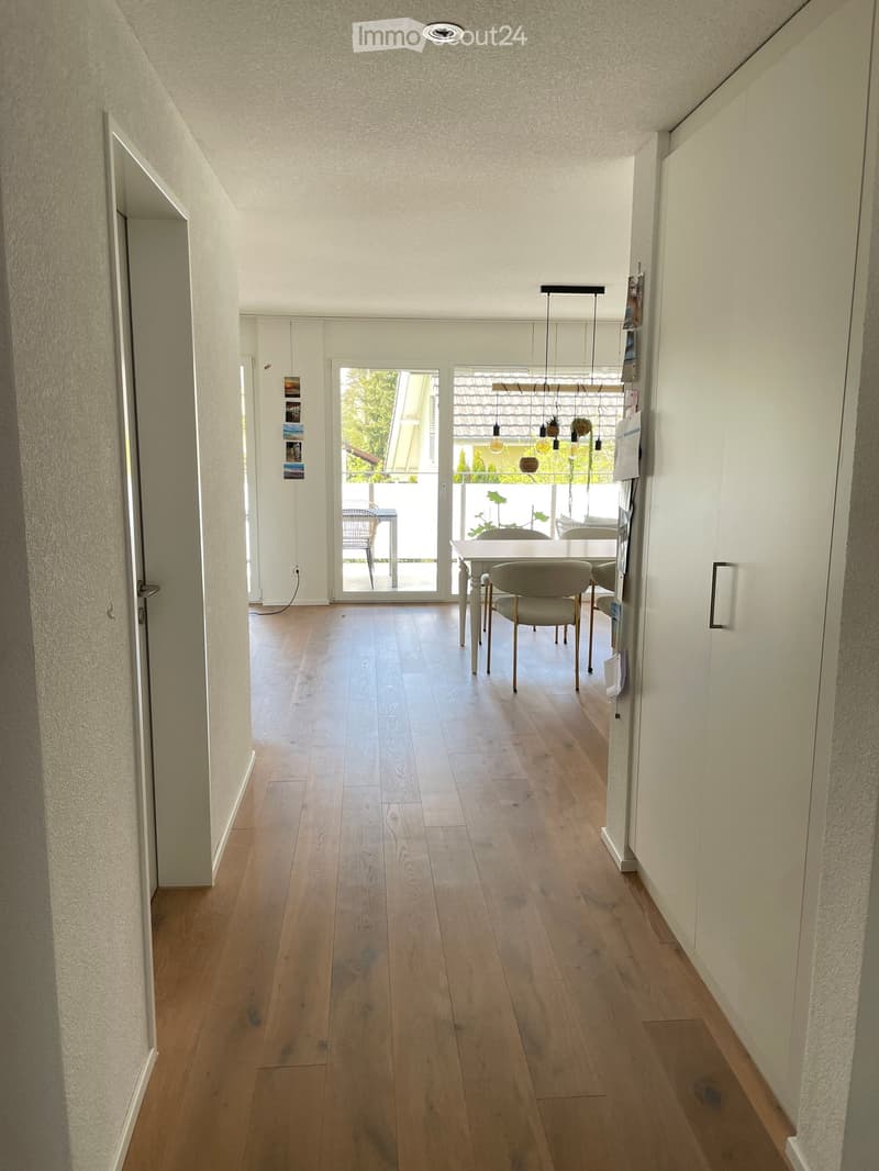 Empty hallway with wooden floor, white walls, glass door leading to a dining area with a table and chairs, and a balcony with plants.