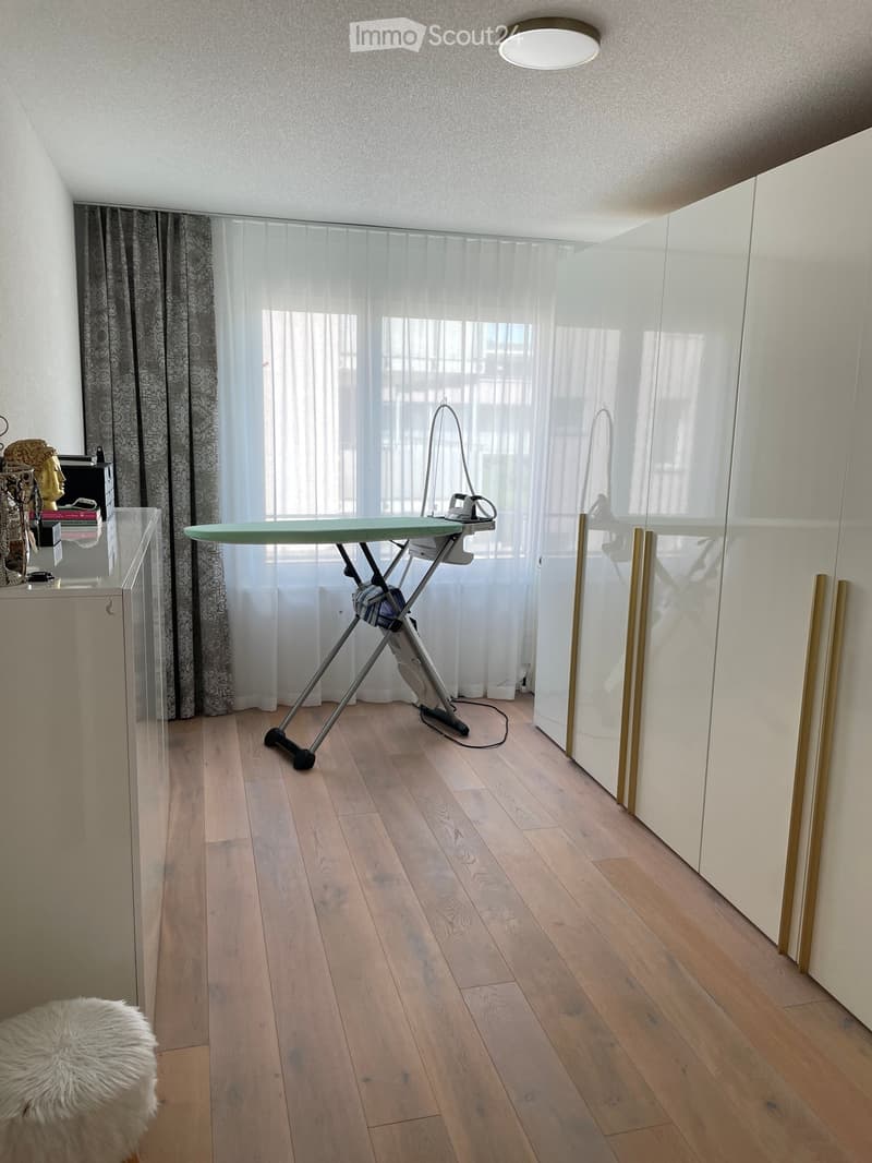 A room with wooden floor, white walls, large windows with white curtains, a white cabinet, a white stool, and an ironing board with an iron on it.