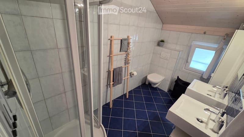 Bathroom. Modern bathroom with blue tiled floor, white tiled walls, a toilet, double sink, towel rack with towels, and a glass-enclosed shower area.