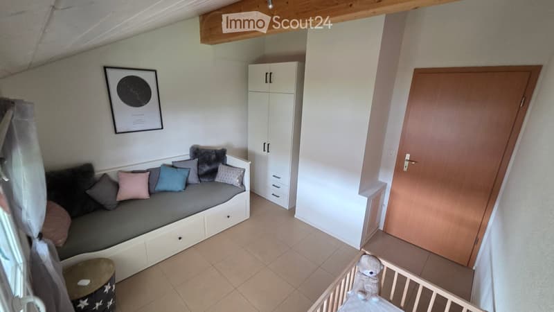 Furnished Room. A cozy, well-lit room with a built-in sofa, colorful pillows, a small round table, a white cabinet, and a baby cradle with a teddy bear toy.