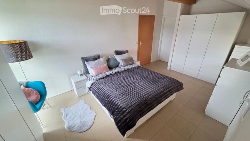 Bedroom. Modern bedroom with a large bed, nightstand, chair, lamp, and wardrobe. The bed is adorned with multiple pillows and a dark blanket. A white rug is placed on the floor near the bed.