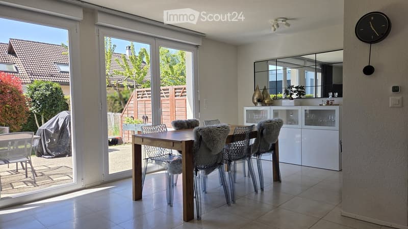 Furnished Room. Bright dining area with a wooden table, clear chairs with fur cushions, white cabinetry, and a large mirror above it. Large sliding glass doors open to a garden.