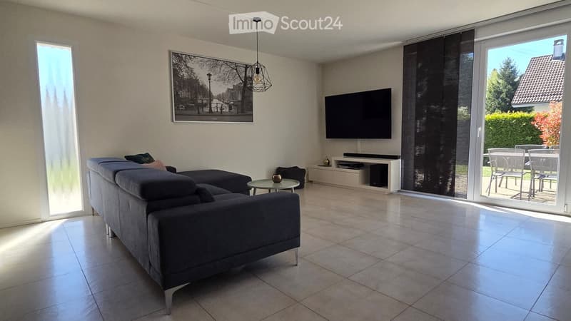 Living Room. Modern living room with a grey sectional sofa, coffee table, flat-screen TV mounted on the wall, and a dining area with a table and chairs visible through the sliding glass doors.