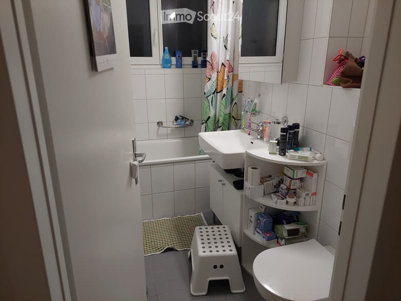 Bathroom. A small bathroom with a white bathtub, sink, toilet, and a shelf holding various toiletries. A shower curtain with a floral design is visible, along with a small white stool and a woven mat on the floor.
