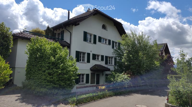 House. Two-story white house with green shutters, multiple windows, and a small balcony on the upper floor. The house is surrounded by lush greenery and trees, with a paved driveway leading to the entrance.