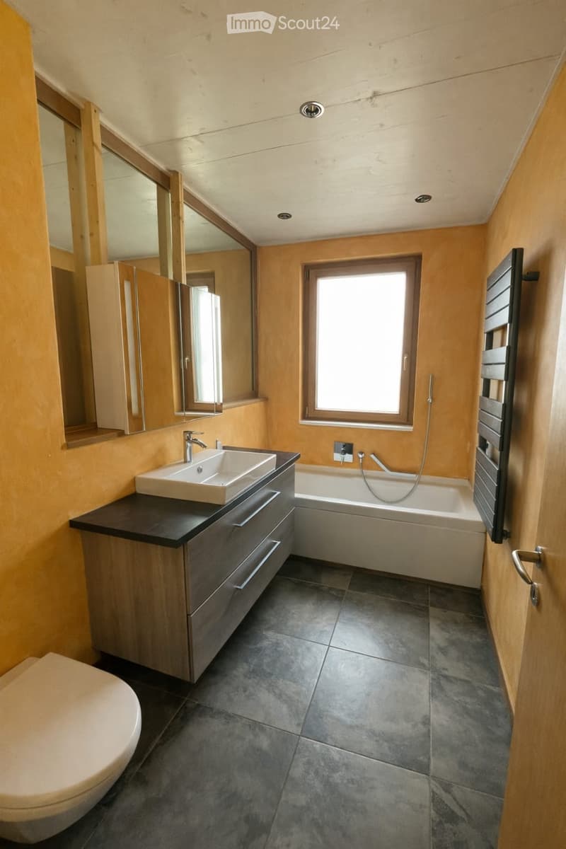 Bathroom. Modern bathroom with a dark grey tiled floor, a white sink with a chrome faucet, a bathtub, a toilet, and a large mirror above the sink. The walls are painted in a warm, light orange color, and there is a window letting in natural light.