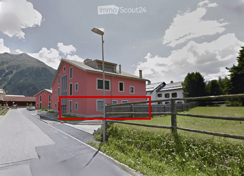 House. A two-story red house with multiple windows and a flat roof, situated in a rural area with a wooden fence and a view of mountains in the background.