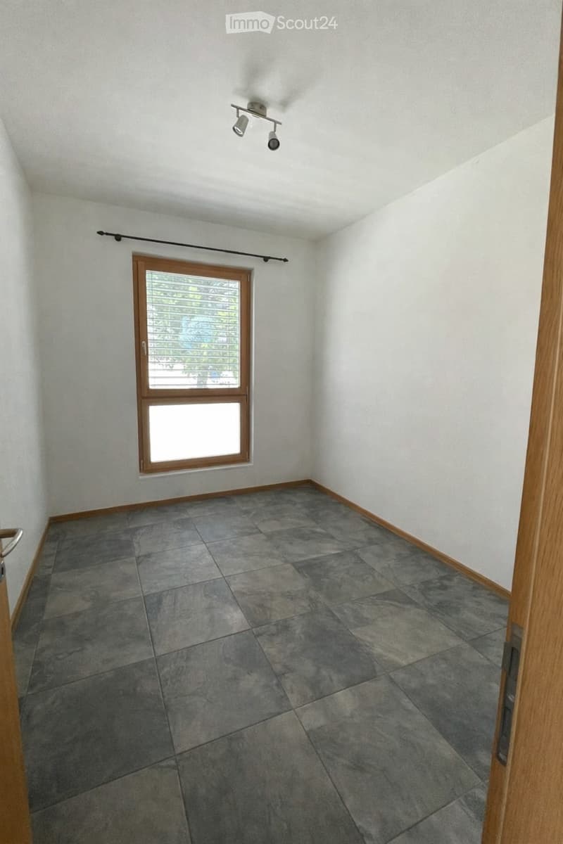 Unfurnished Room. Empty room with grey tiled floor, white walls, and a window with wooden frame and blinds. Ceiling light fixture visible.