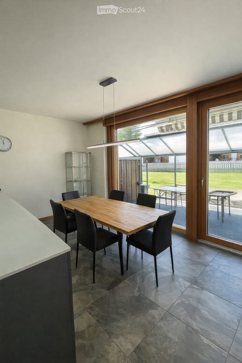Furnished Room. Dining room with a wooden dining table, six black chairs, a glass cabinet, and a clock on the wall. Large sliding glass doors lead to an outdoor patio.