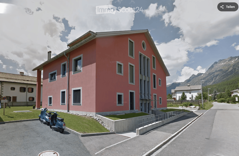 Building. A two-story red building with multiple windows and a flat roof, situated in a scenic mountainous area with a paved driveway and scooters parked nearby.