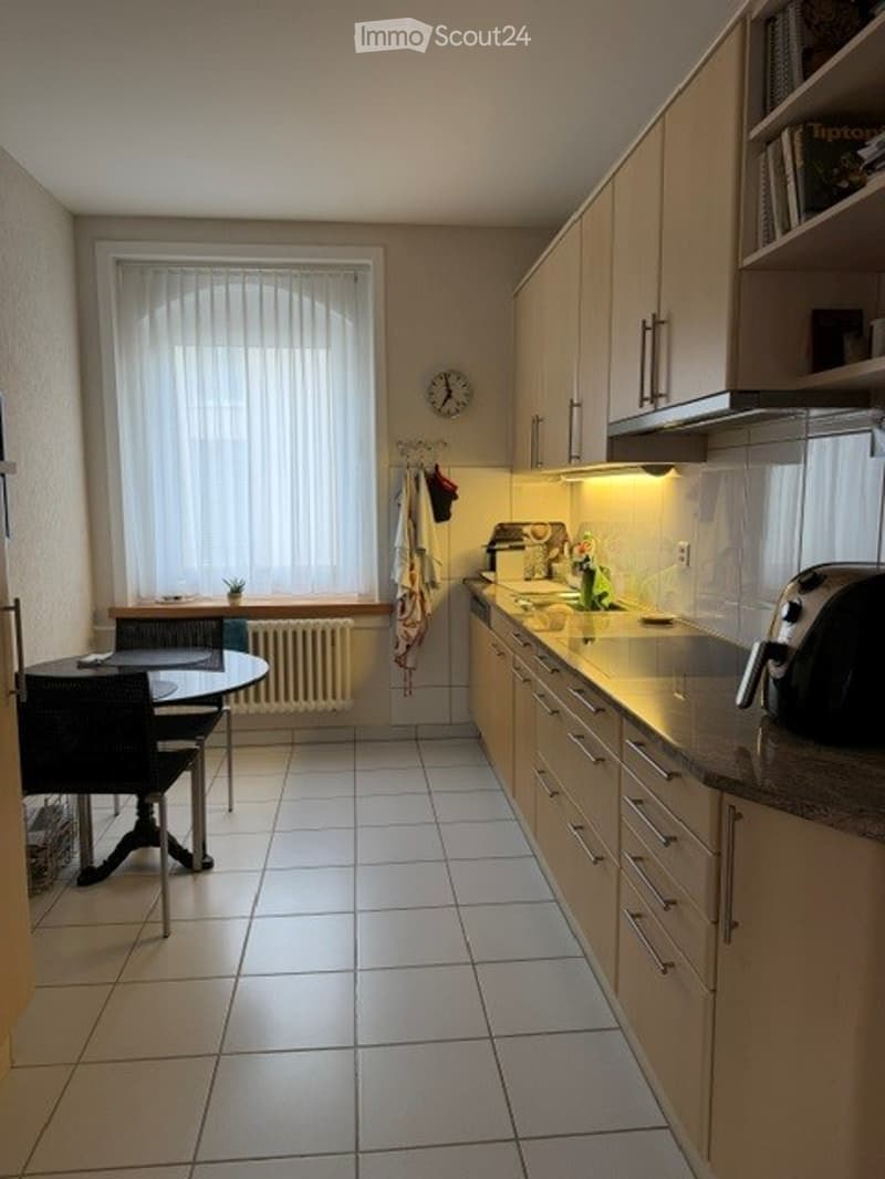 Kitchen. Bright kitchen with white tiled flooring, light wood cabinets, a central island with sink and faucet, and a small dining area with a table and chairs. A window with blinds provides natural light, and a clock is mounted on the wall.