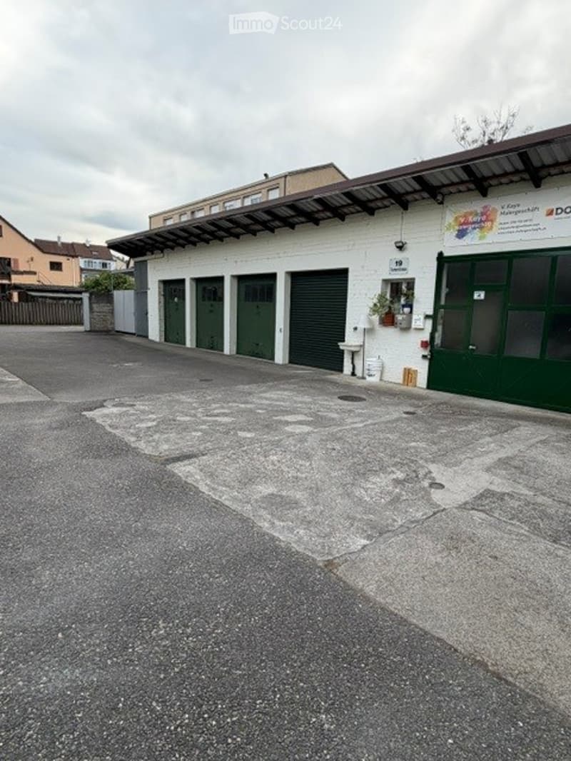 Building. A white building with multiple garage doors and a covered entrance area. The ground is paved, and there is a small garden area near the entrance.