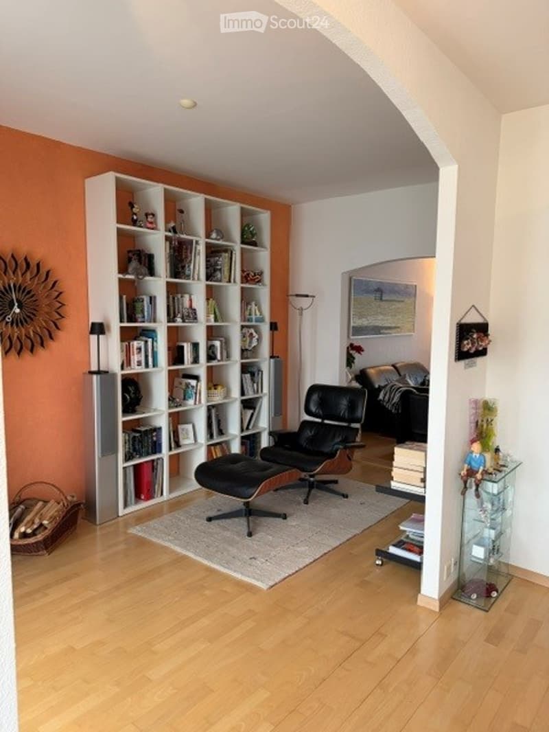 Furnished Room. Modern living room with light wooden flooring, white built-in bookshelf filled with books and decorative items, black leather sofa and ottoman, floor lamp, and a glass cabinet with various items.