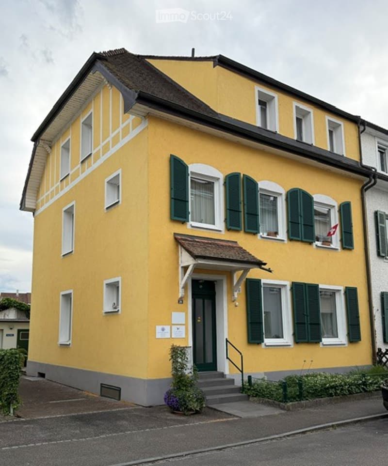 House. Two-story yellow house with white trim, green shutters, and a small porch with a flag. The entrance has a staircase leading to a green door.