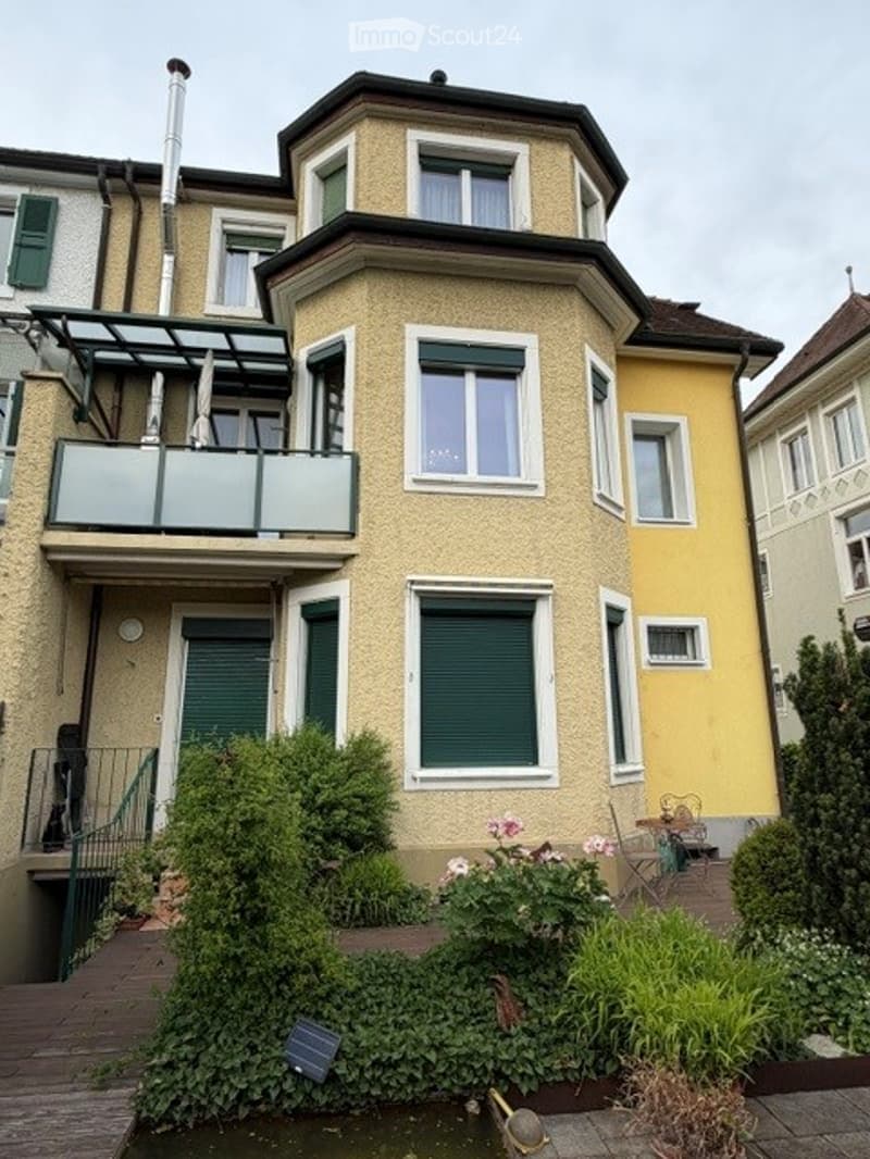 Building. Multi-story residential building with a balcony, green shutters, and a small garden area with plants and a small pond.