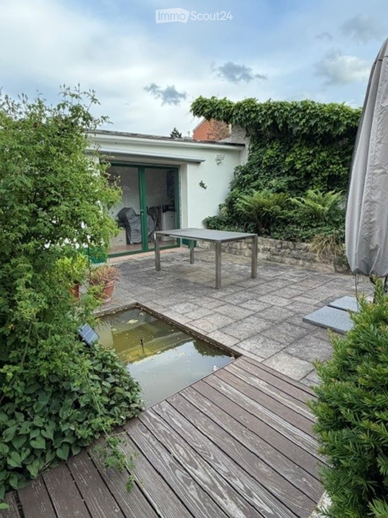 Garden / Outdoor Area. A well-maintained garden patio with a small pond, wooden decking, a metal table and chairs, surrounded by lush greenery and a covered outdoor area.