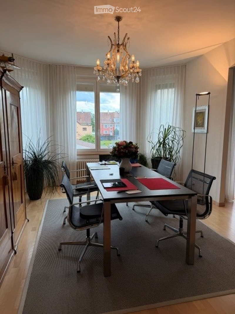 Furnished Room. Brightly lit room with large windows, a long conference table surrounded by chairs, a chandelier hanging from the ceiling, and potted plants near the windows.