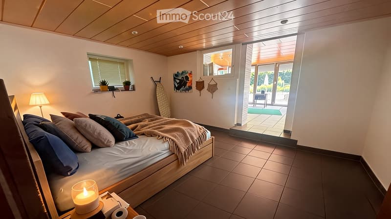 Bedroom. Cozy bedroom with a wooden bed, multiple pillows, a blanket, a table lamp, a candle on a nightstand, a mirror, art on the wall, a surfboard leaning against the wall, and a sliding glass door leading to a balcony or terrace area with outdoor furni