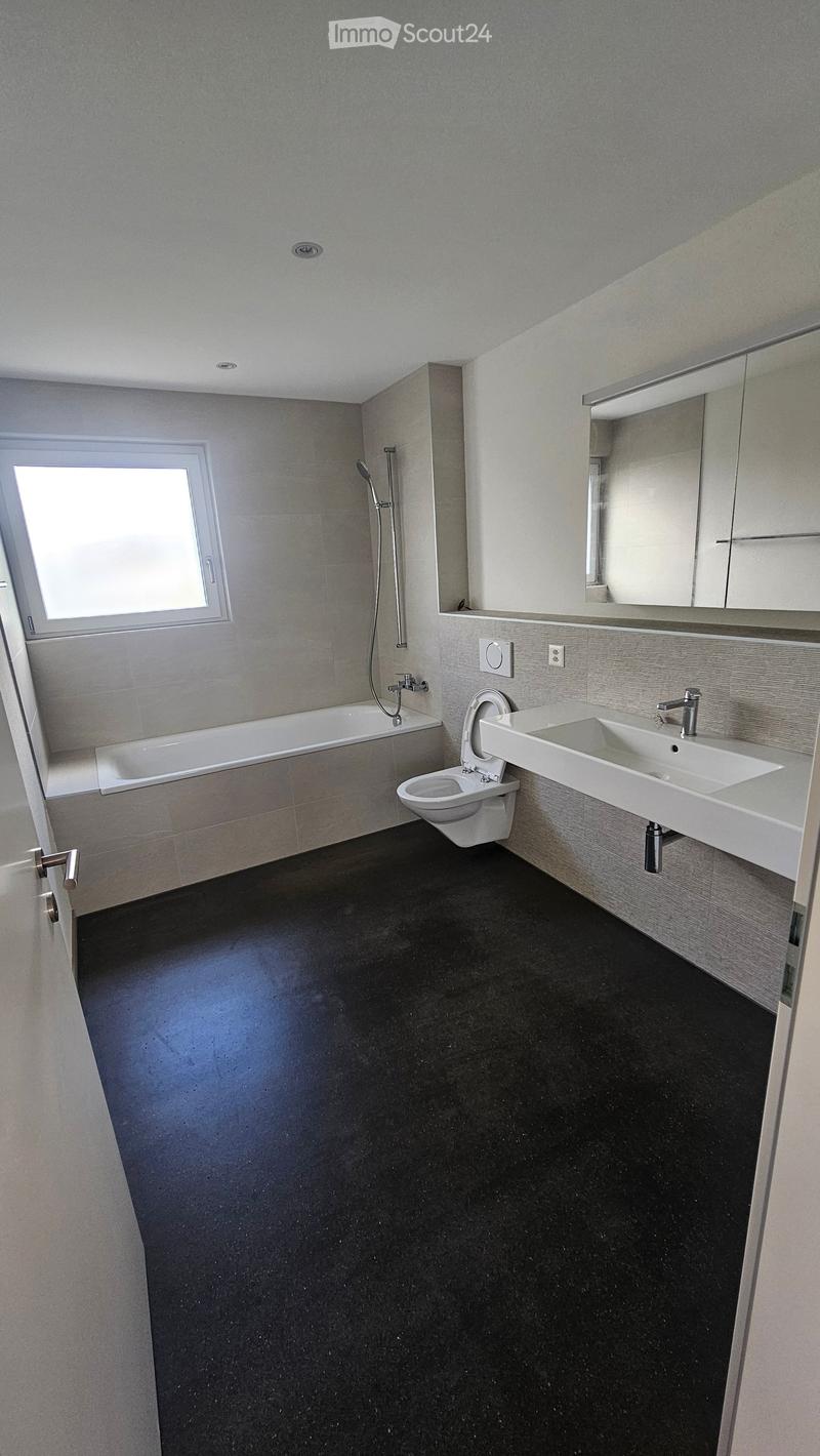 Bathroom. Modern bathroom with a bathtub, toilet, sink, and large mirror. Features a dark floor and grey tiled walls.
